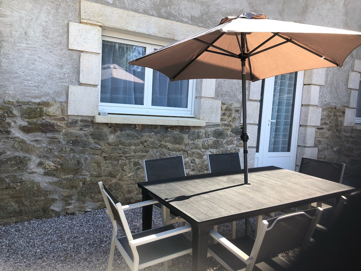 An outdoor dining area is presented, featuring a large black table surrounded by six chairs. A sizable umbrella provides shade, and a stone wall compliments the setting. A window can be seen in the background, enhancing the space's overall charm.
