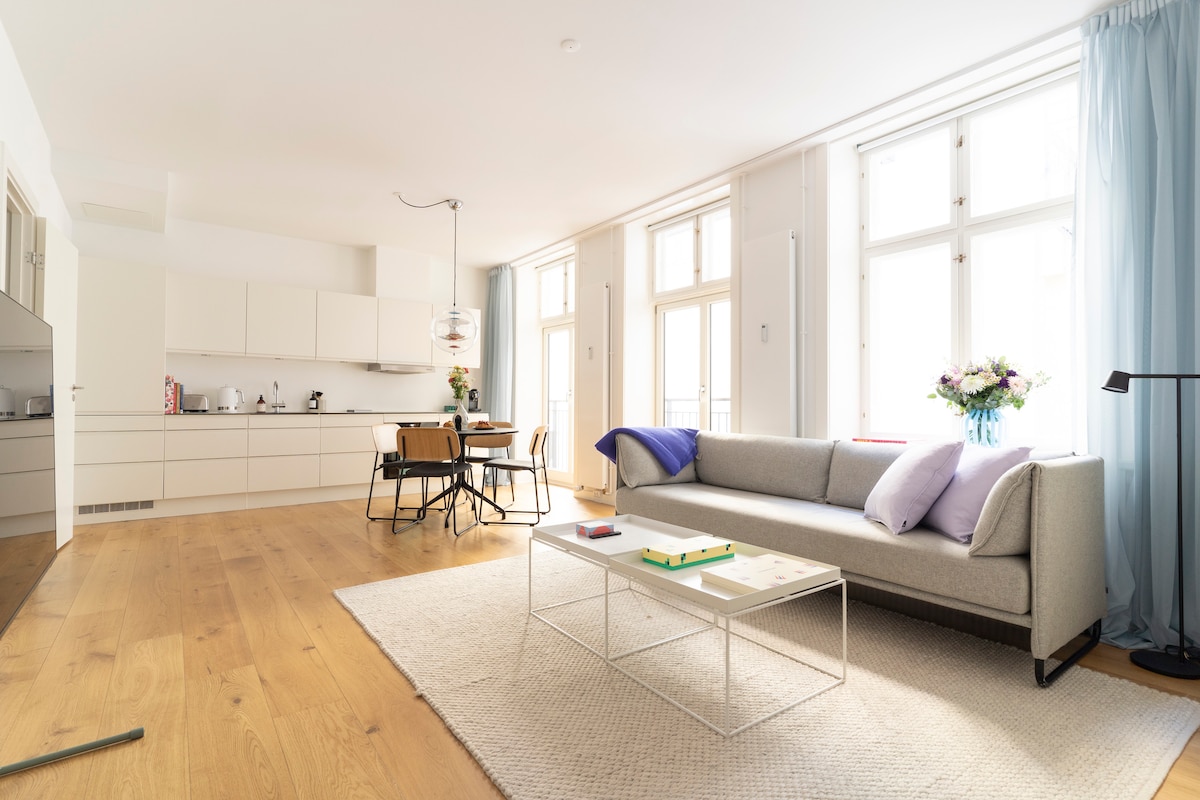 An open living area features a light-colored sofa with cushions, positioned near a coffee table on a textured rug. Large windows offer ample natural light, while a dining table and chairs are visible in the background next to a modern kitchen.