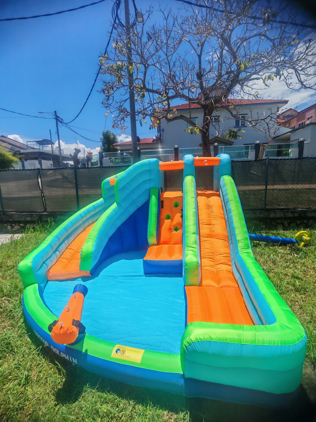 An inflatable water slide and pool set is situated on a grassy area, featuring bright green and orange colors. The slide has two distinct sections for sliding and climbing, with some colorful climbing balls integrated into the design. A clear sky is visible in the background.