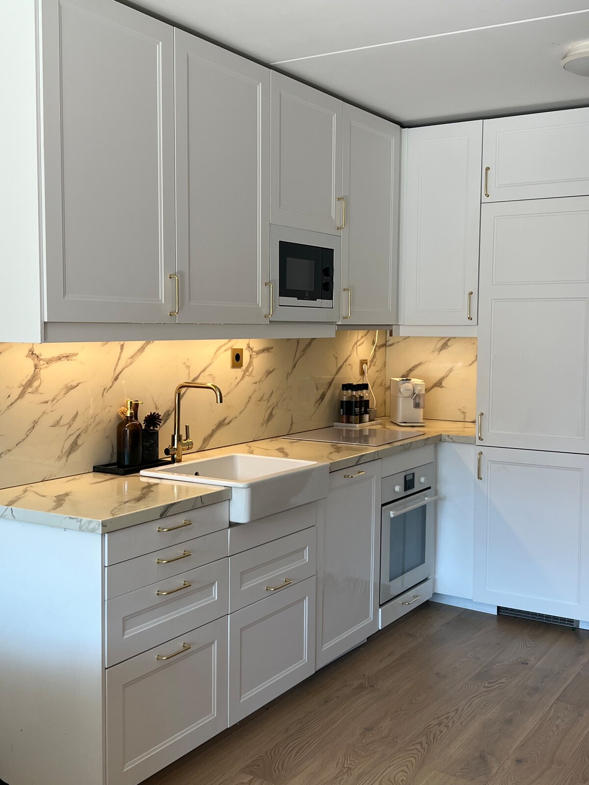 A modern kitchen is presented, featuring sleek white cabinetry with gold hardware. A large farmhouse sink is situated beneath a wall-mounted microwave. The marble backsplash adds a textured element, while stainless steel appliances and a coffee maker are visible, completing this functional and stylish space.