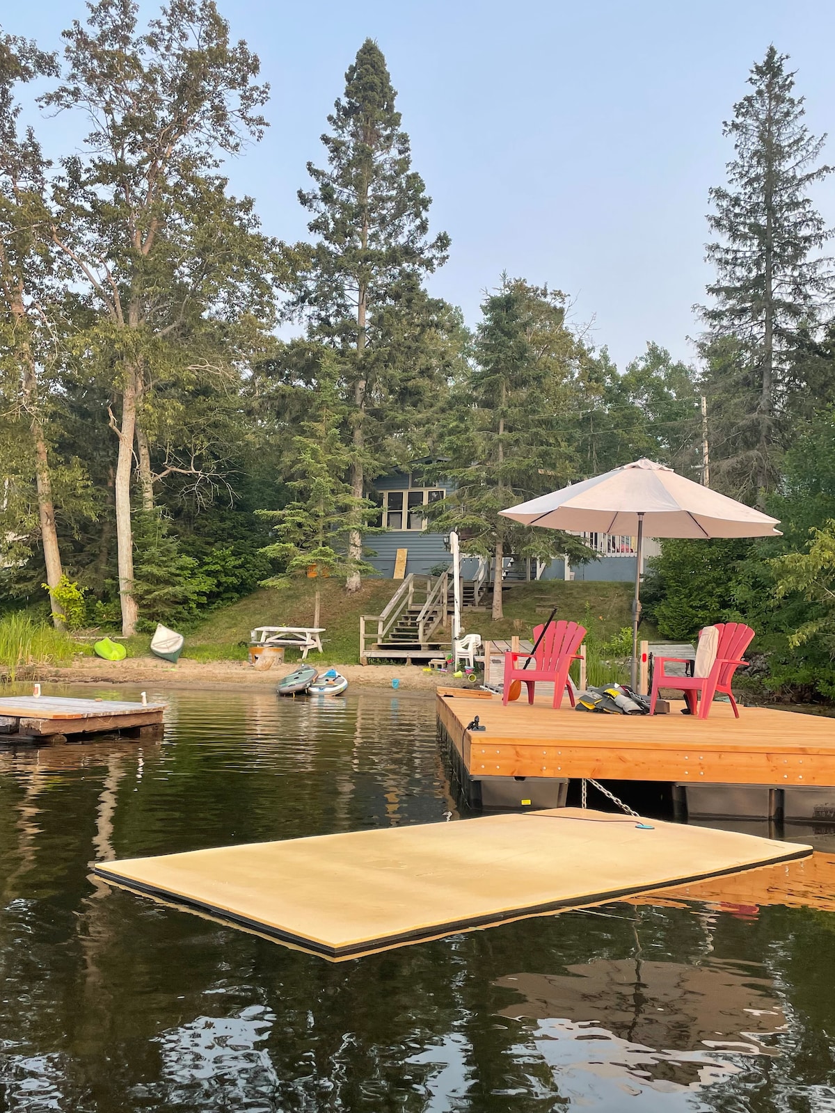 A wooden dock extends into calm waters, featuring two red chairs and an umbrella providing shade. The cottage is visible in the background, surrounded by tall trees. Various water toys and equipment are stationed nearby, emphasizing a natural and inviting lakeside setting.