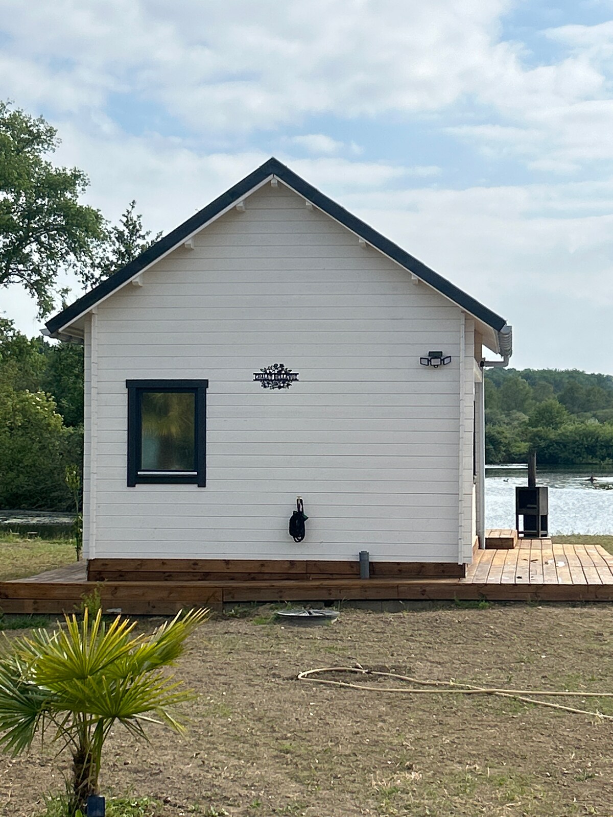 The exterior of a quaint white chalet is depicted, showcasing simple architectural lines. A small wooden deck extends from the back, bordering a body of water. A single window is present on the side, allowing light into the interior space.