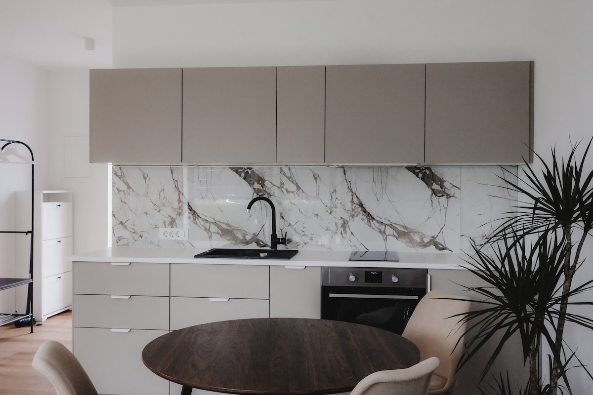A modern kitchen features sleek cabinetry in soft beige tones, complemented by a striking marble backsplash. A black faucet is positioned above the stove, and a circular wooden dining table is visible alongside pale accent chairs. Natural light creates a bright atmosphere.
