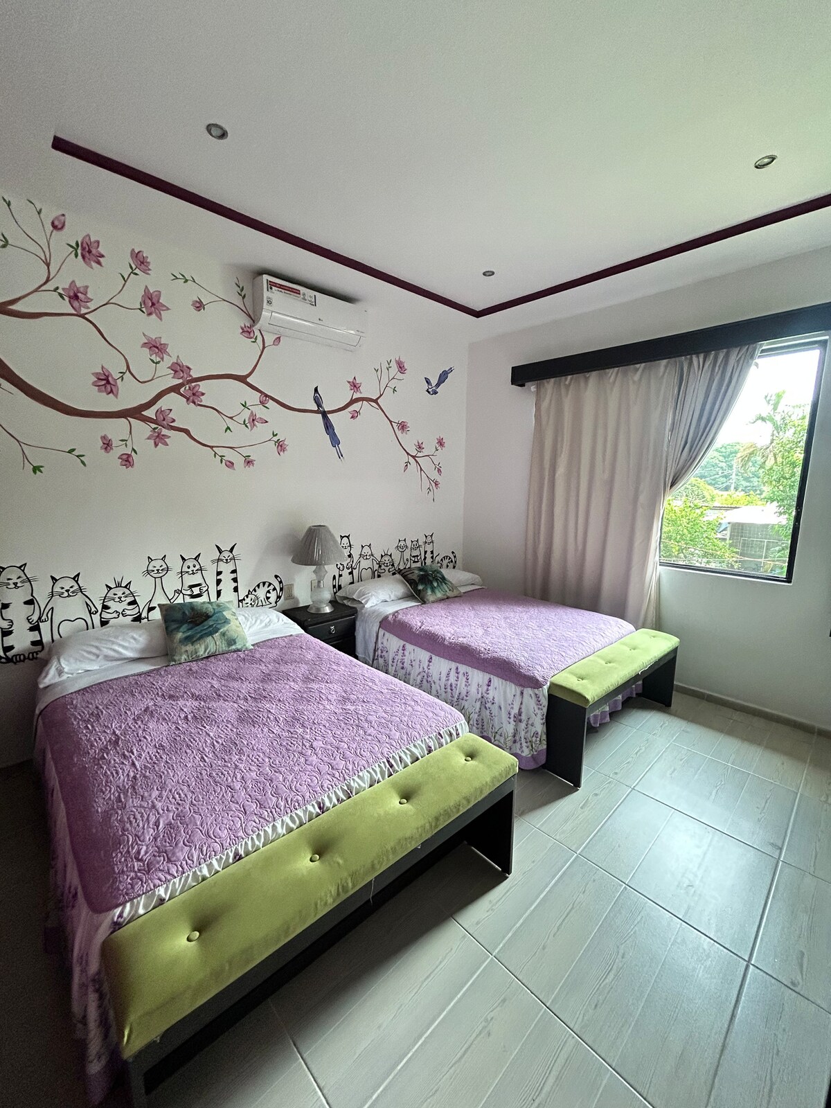 A bright sleeping area is highlighted by two spacious beds covered in lavender bedspreads. A mural featuring birds and trees adorns the wall, complemented by a bedside lamp. Natural light filters through a window covered by light curtains, enhancing the room's airy feel.