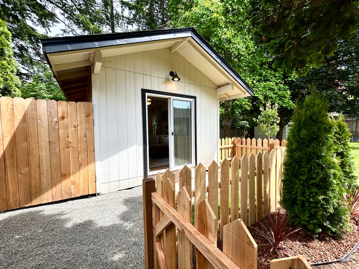 A private entrance is framed by a fenced pathway leading to a charming casita. The exterior features crisp white siding and a sloped roof. Lush greenery surrounds the space, enhancing the sense of seclusion and tranquility.