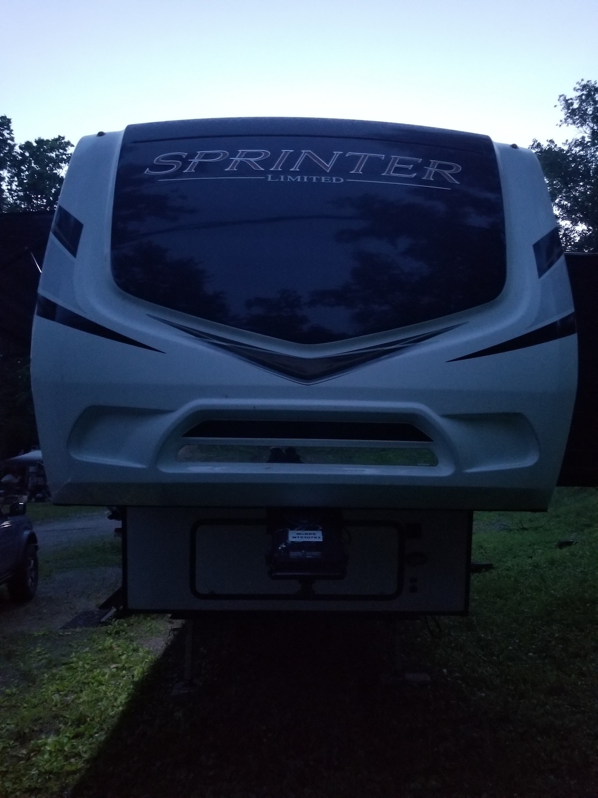 The RV is showcased in soft evening light, highlighting its sleek exterior design. The prominent logo is situated at the top, while the overall structure reveals large front windows and a distinct aerodynamic shape. The surroundings include greenery, suggesting a natural setting.