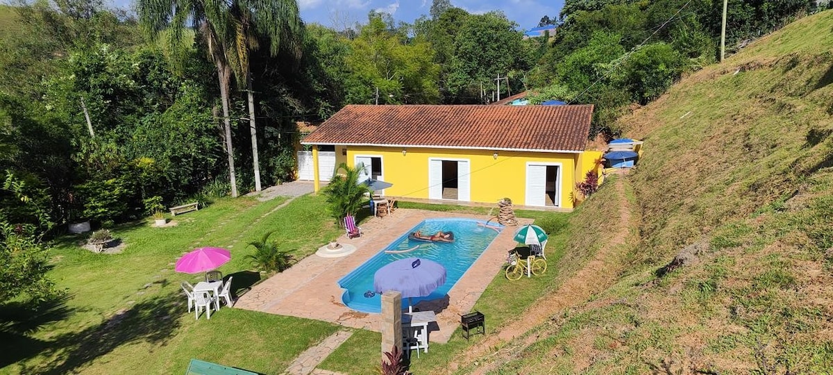 A colorful single-story house with a yellow exterior is surrounded by lush greenery. The outdoor area features a swimming pool with shade provided by umbrellas and lounge chairs. A garden space is present with a seating arrangement, enhancing the tranquil setting.