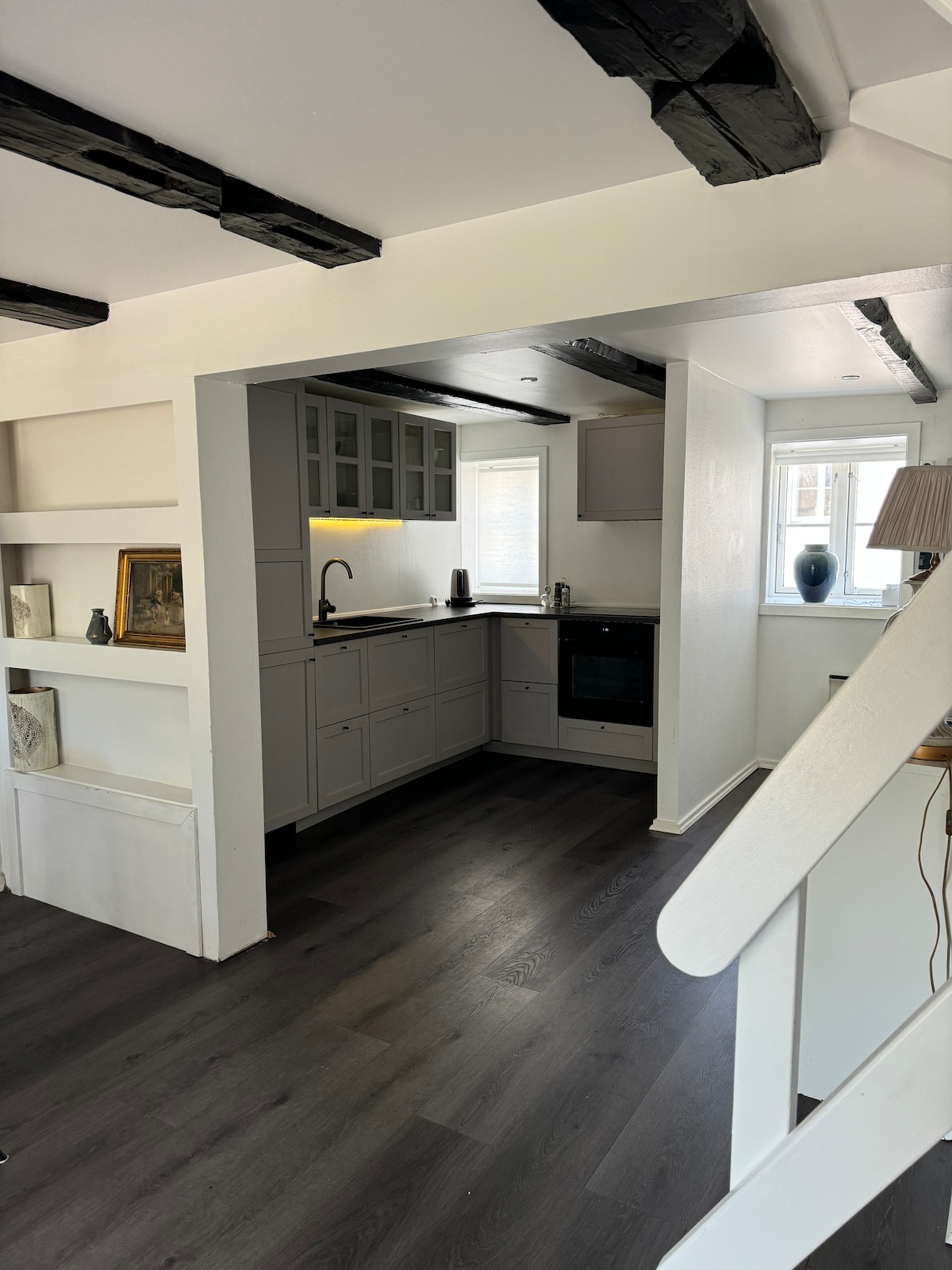The kitchen area features modern cabinetry complemented by sleek countertops. A window provides natural light, illuminating the cooking space and the stovetop. A staircase is visible, leading to the upper level. Decorative framed artwork is arranged on adjoining shelves.
