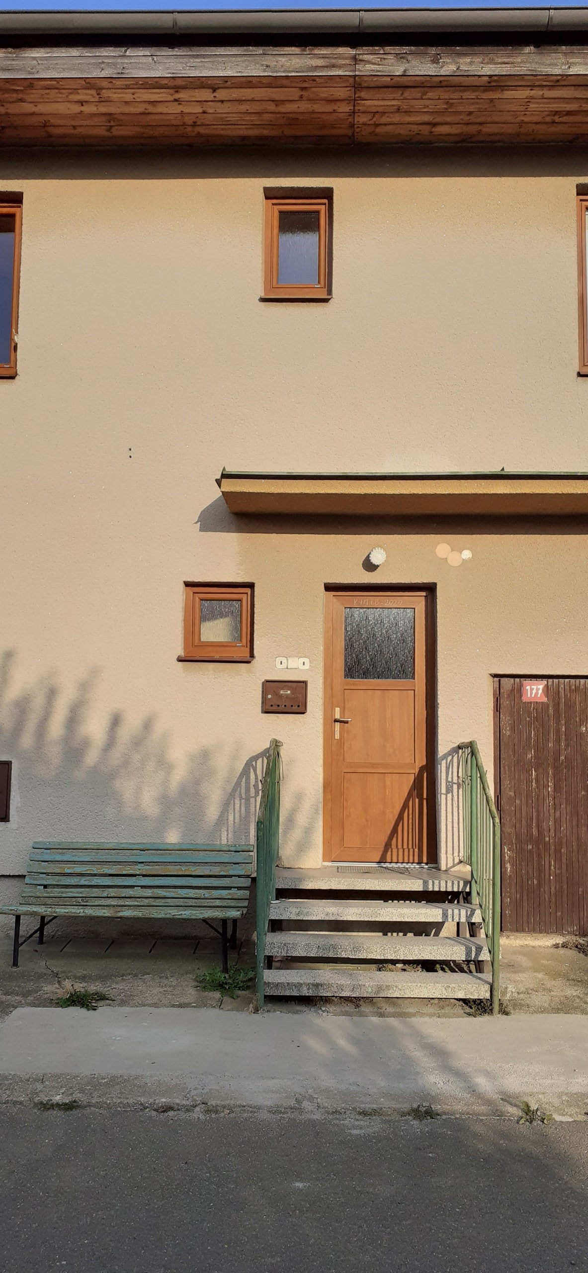 An inviting exterior view of a two-story building is shown. A set of steps leads to a wooden door framed by two small windows. A bench is positioned to the side, and a wooden panel marks the entrance path.