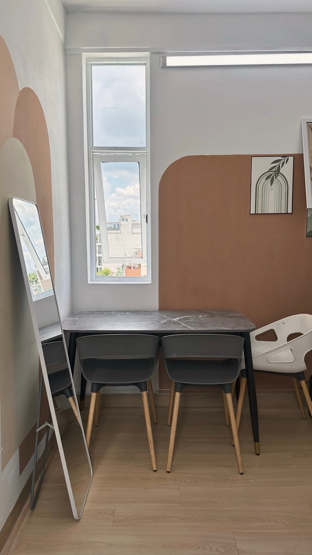 A small dining area features a triangular marble table surrounded by three sleek grey chairs and one white chair. A full-length mirror is positioned nearby, with natural light filtering through two windows that offer views of the outside.