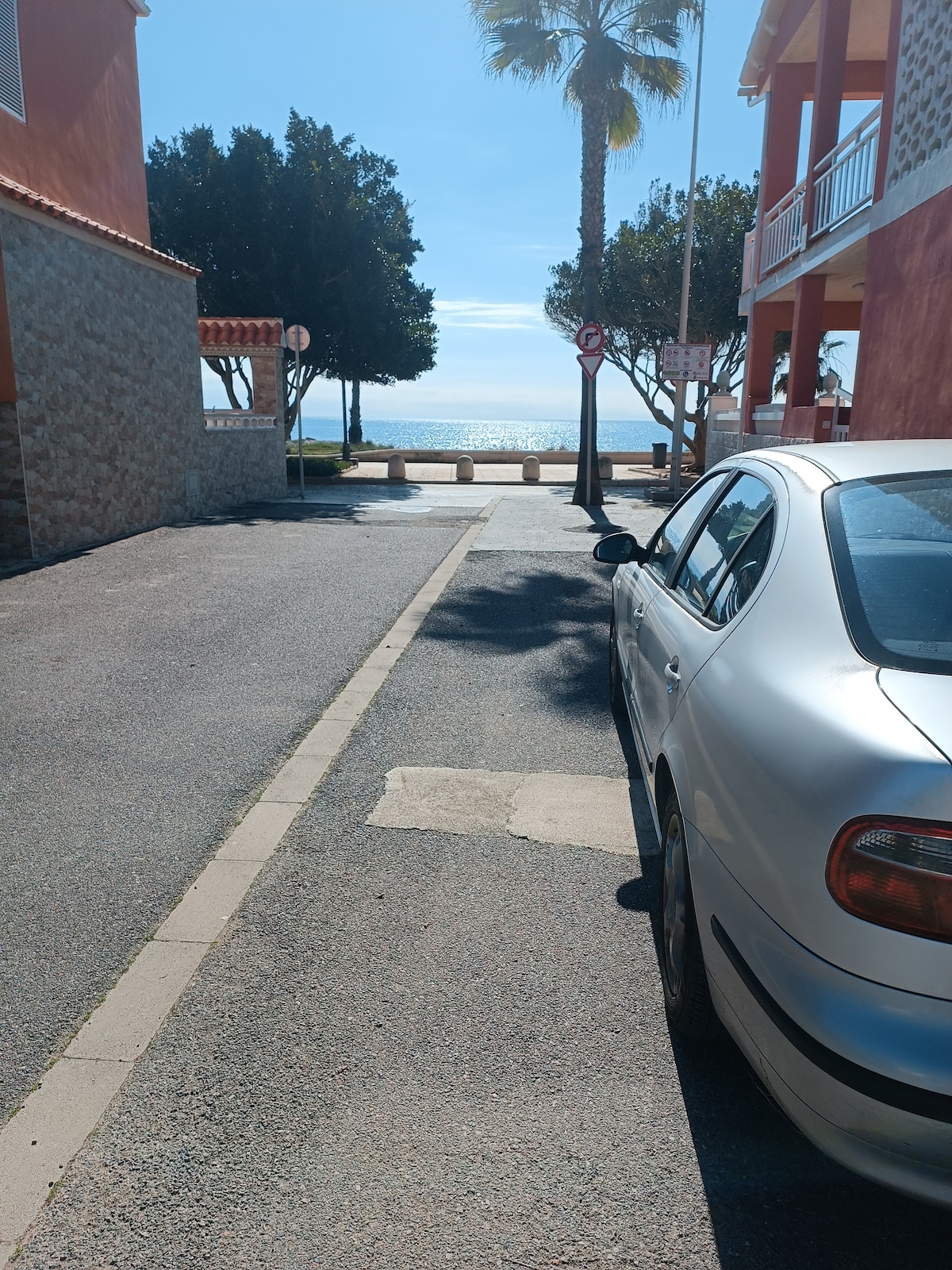 A narrow street is depicted, leading towards the shimmering sea at a distance. A silver car is parked alongside the curb, with palm trees and buildings visible on either side. Clear blue skies enhance the serene atmosphere.