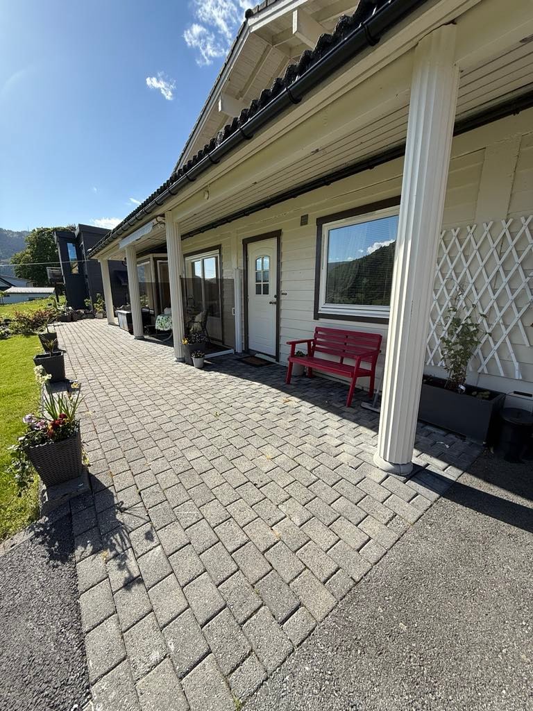 A welcoming outdoor area features a paved pathway lined with flower pots. The white facade of the building is complemented by two entrance doors, framed by decorative columns. A red bench provides a resting spot while the bright sky above enhances the peaceful setting.