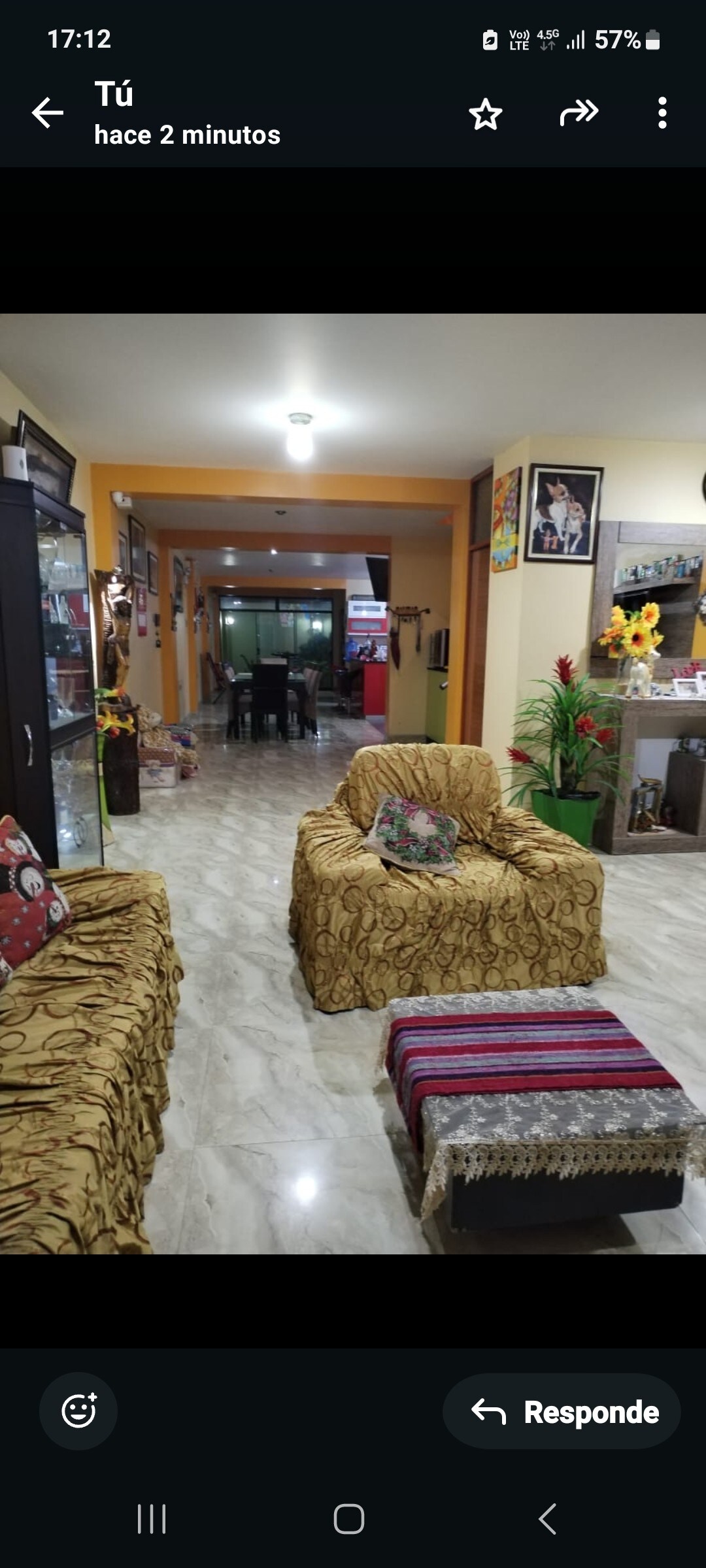 A welcoming living area is furnished with two patterned sofas and a small, colorful table at the center. The space showcases tiled flooring and is illuminated by overhead lighting. In the background, a dining area is visible, featuring a table and chairs. Decorative elements are observed throughout.