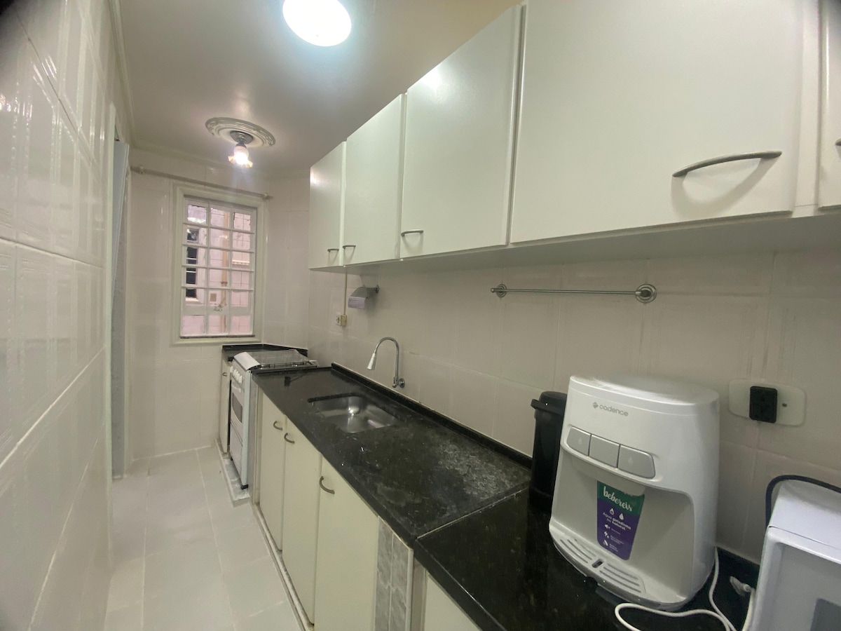 A streamlined kitchen is presented, featuring light-colored cabinets and a dark granite countertop. Essential appliances, including an oven, sink, and a compact coffee maker, are visible. Natural light enters through a nearby window, highlighting the modern fixtures and clean tilework.