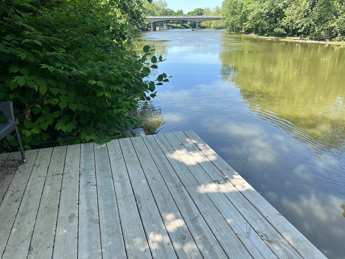 A wooden dock extends over calm waters, surrounded by lush greenery. The river reflects gentle ripples, and a bridge is visible in the distance. A chair is positioned on the dock, providing a serene view of the waterway.