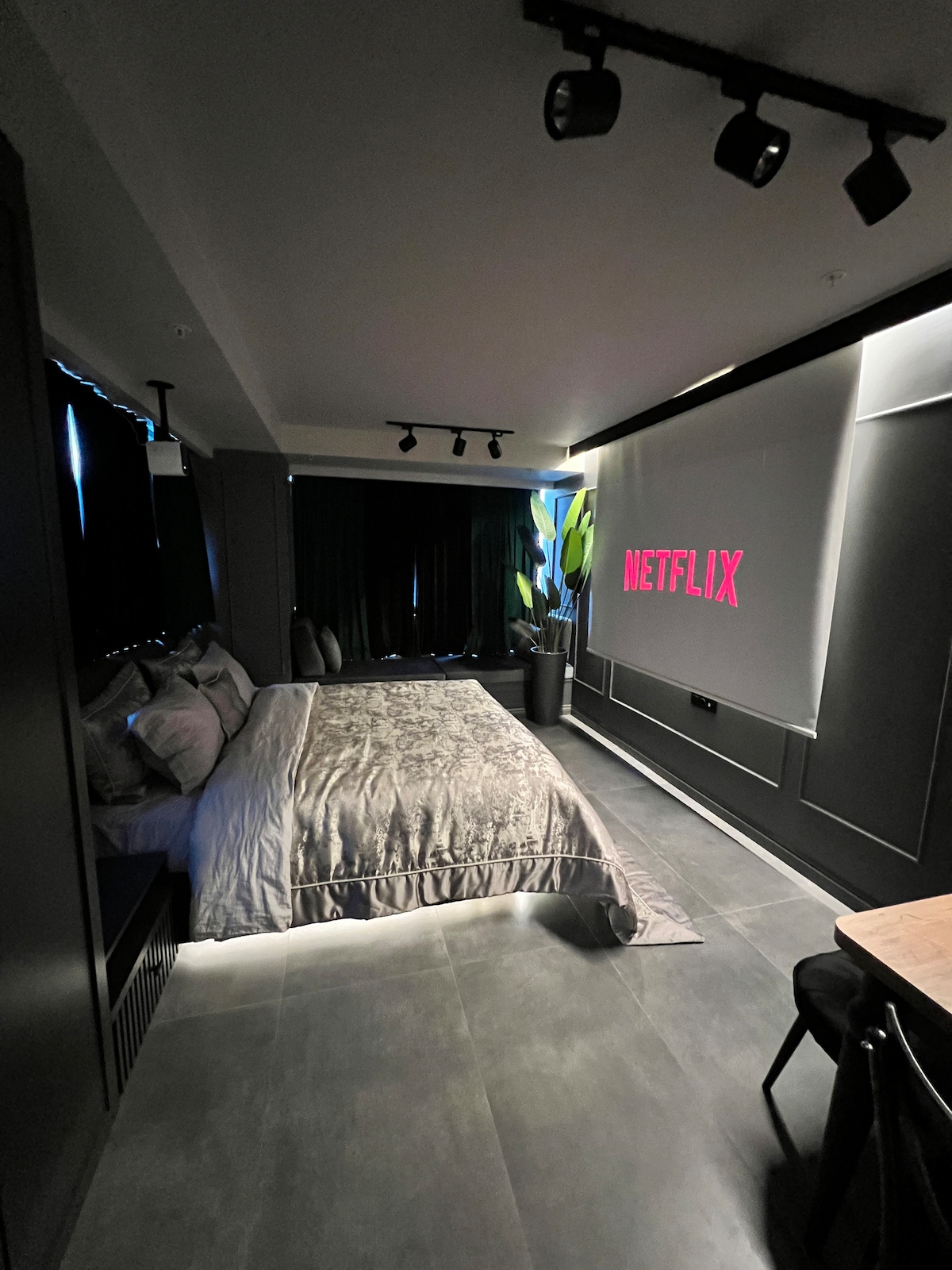 A sleek bedroom features a spacious bed with soft bedding, accompanied by a large screen displaying the Netflix logo. Dark curtains frame the window, while subtle illumination from spotlights enhances the modern aesthetic of the space.