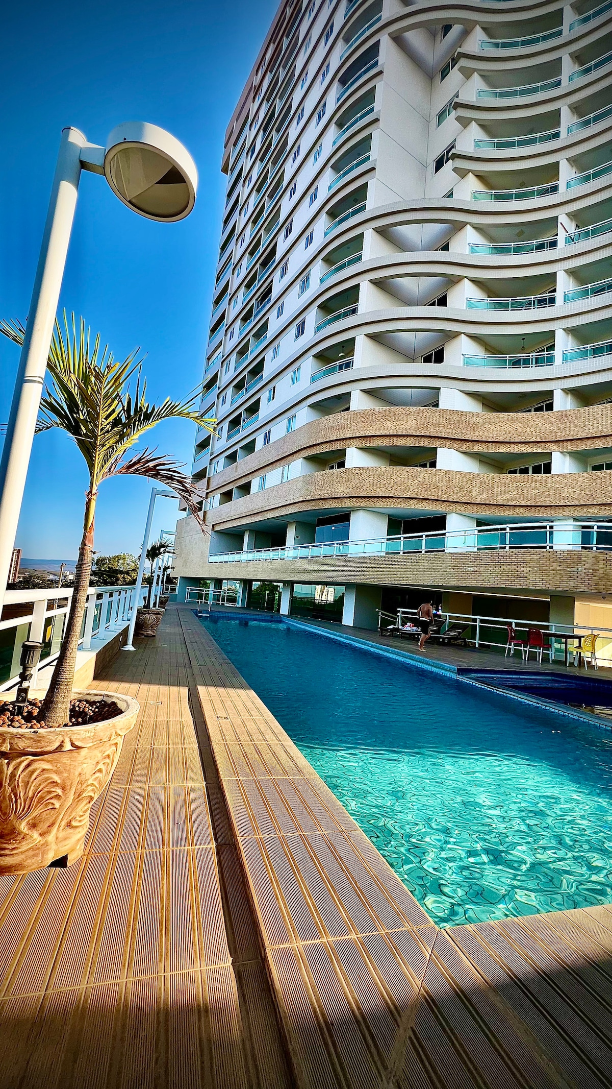 A serene poolside area is showcased, featuring a modern building in the background. The pool reflects the blue sky, with lounge seating available nearby. Potted palm plants add a touch of greenery to the space, enhancing the overall relaxation environment.