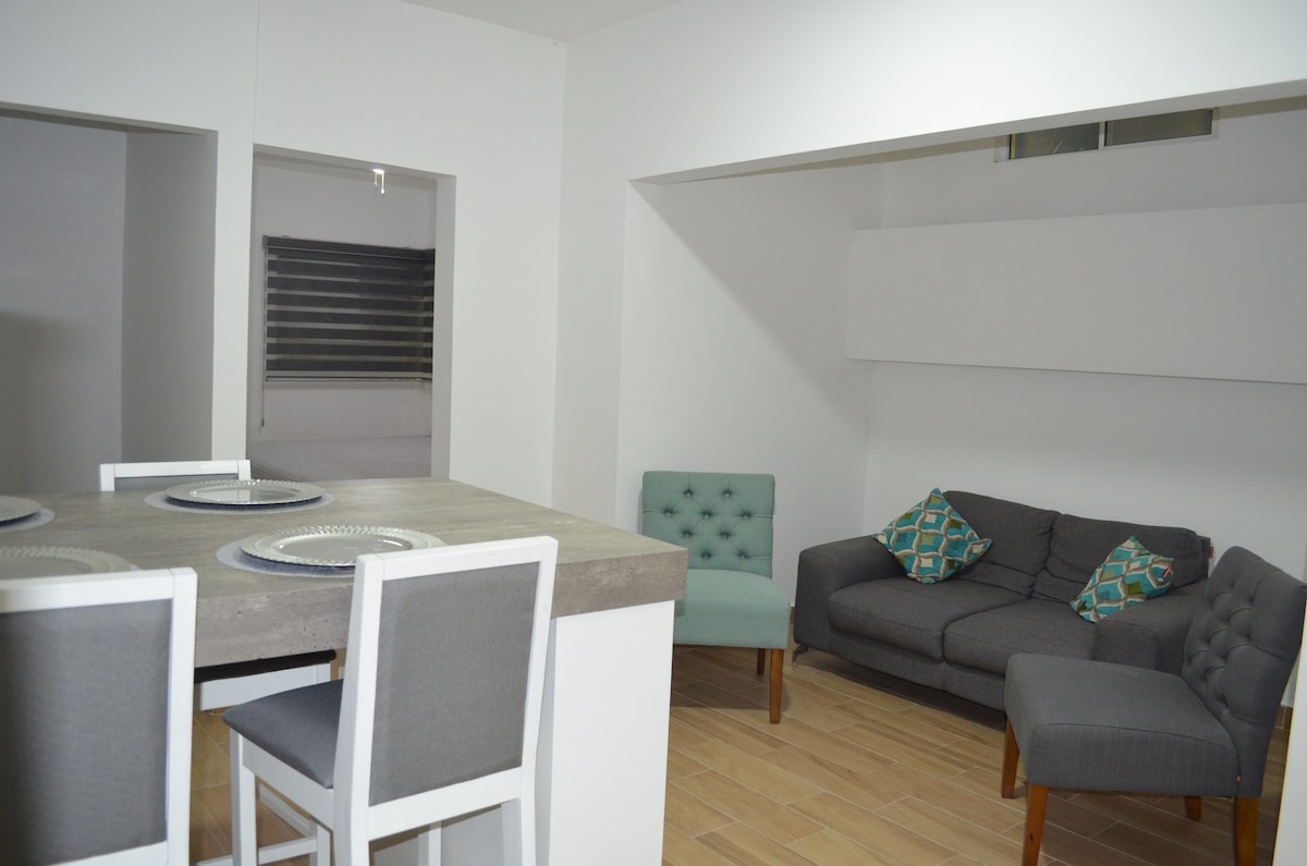 An open living area features a gray sectional sofa adorned with patterned cushions. A light wooden dining table is set with placemats, accompanied by four upholstered chairs. Natural light enters through a window with blinds, enhancing the minimalist decor.