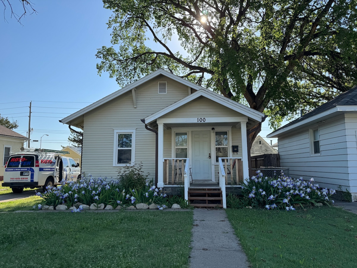 A single-story house is showcased with a light-colored exterior. A welcoming front porch with white railings leads to the entrance. The yard features well-maintained green grass and blooming purple flowers, providing a pleasant view. A large tree offers shade to the front area.