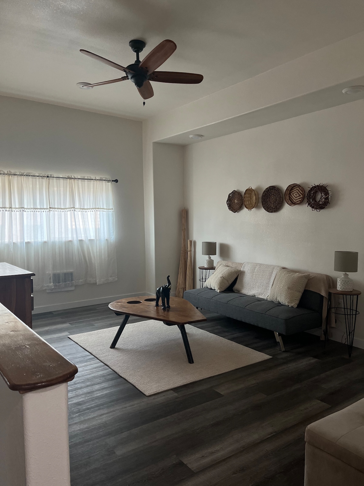 A living area features a comfortable futon centered on a light rug, accompanied by a round wooden coffee table. Soft lighting is provided by two lamps on either side. Natural light filters through the sheer curtains, and woven decor adorns the wall.