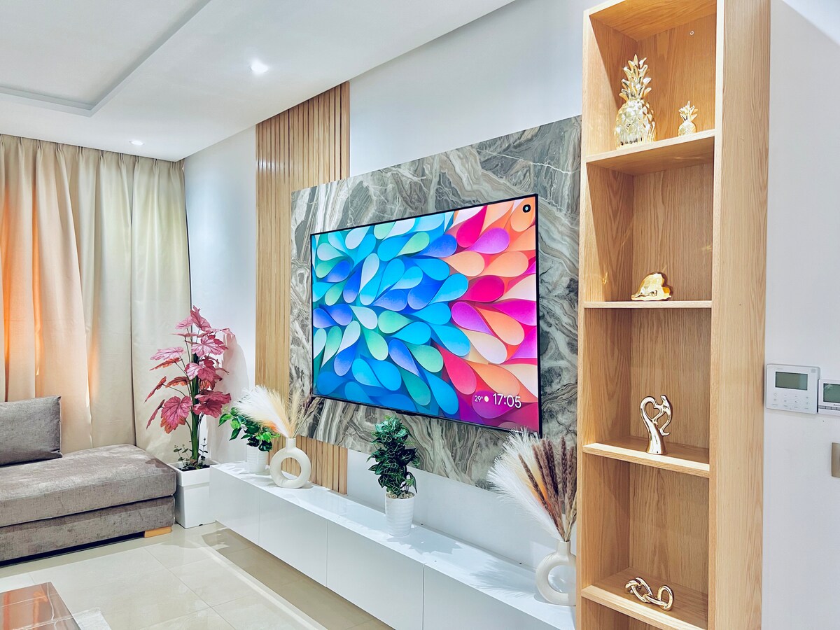 A modern living area is presented with a large flat-screen television mounted on a stylish stone wall. Beneath the TV, a sleek white console holds decorative items. Natural light filters through sheer curtains, enhancing the warm tones of the surrounding furnishings.