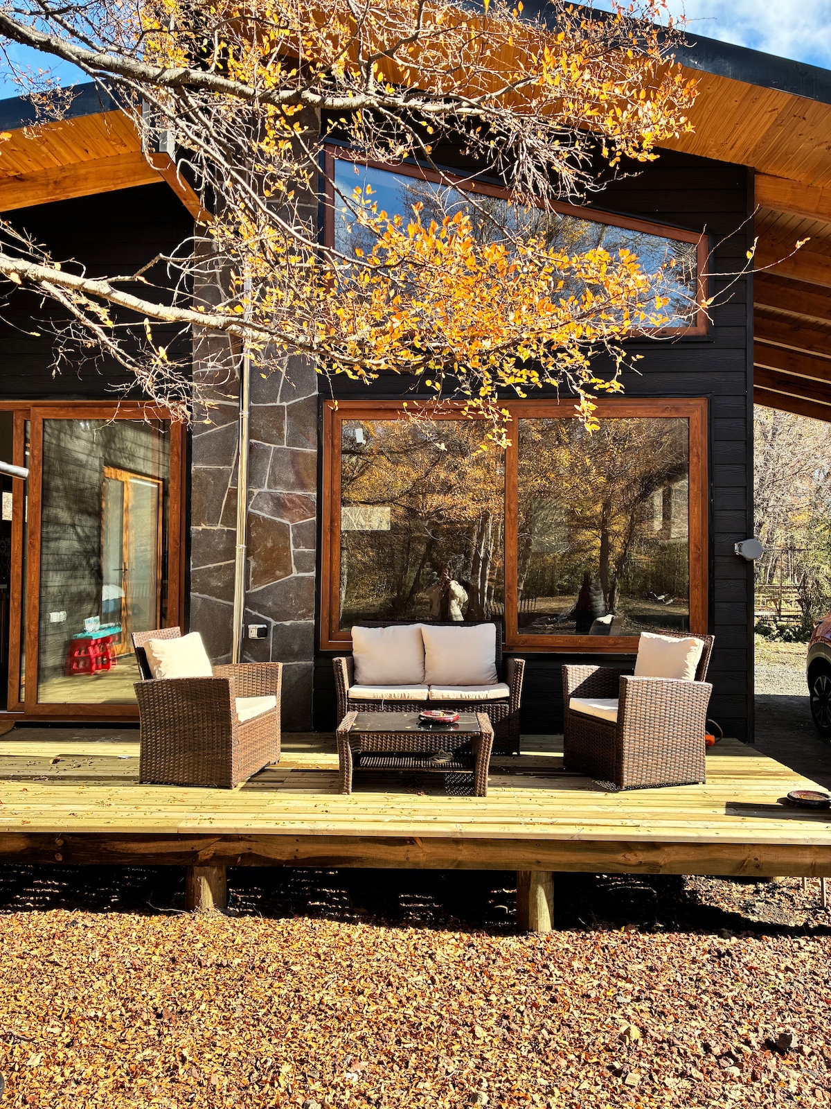 An outdoor seating area features two cushioned chairs and a coffee table on a wooden deck. The space is framed by large windows reflecting the surrounding trees with autumn leaves, enhancing the connection to nature.