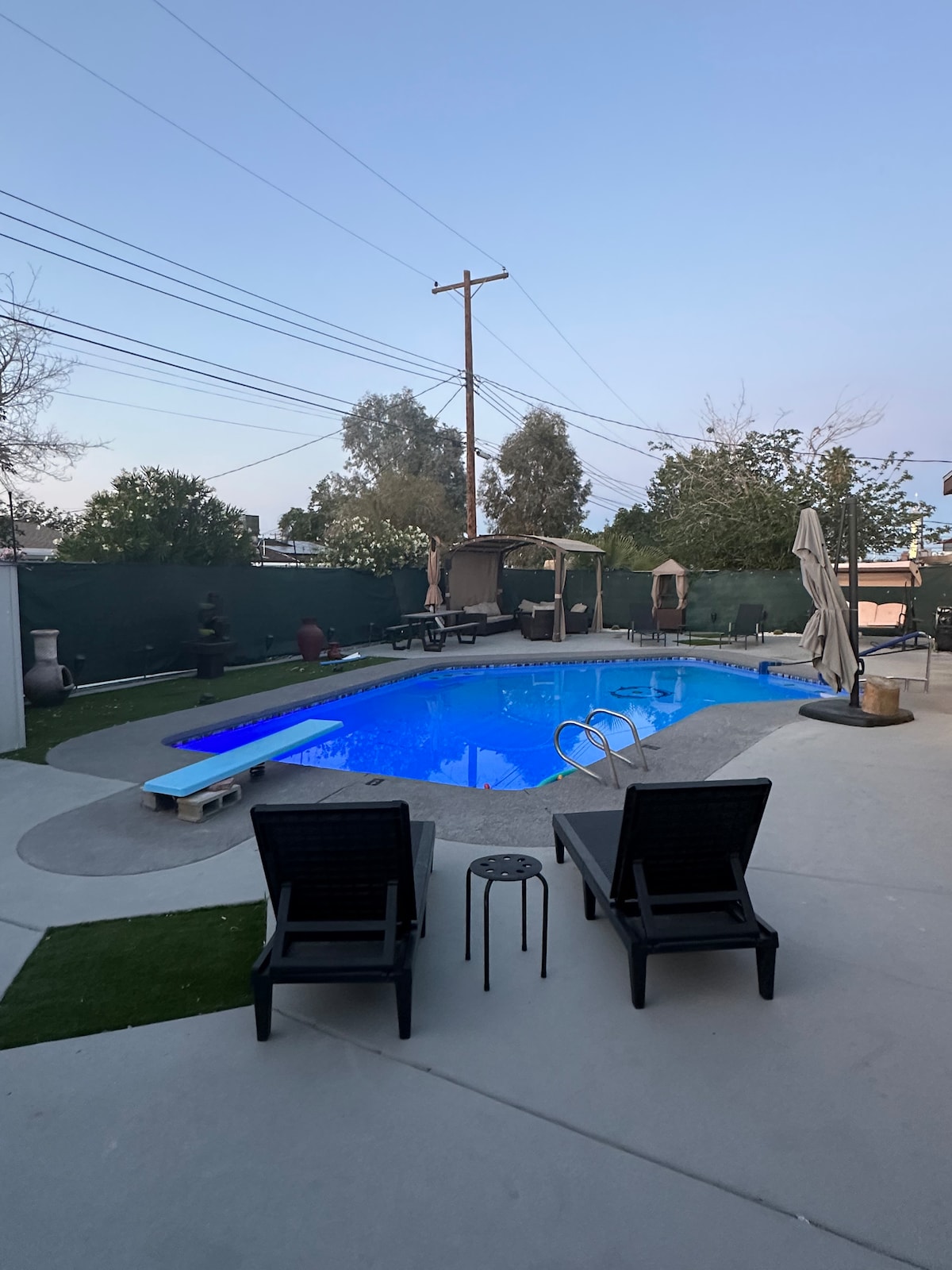 An outdoor space features a swimming pool with bright blue water, surrounded by a paved patio. Two black lounge chairs are positioned near the pool, along with a small side table. Lush greenery and a canopy provide privacy and a sense of tranquility.