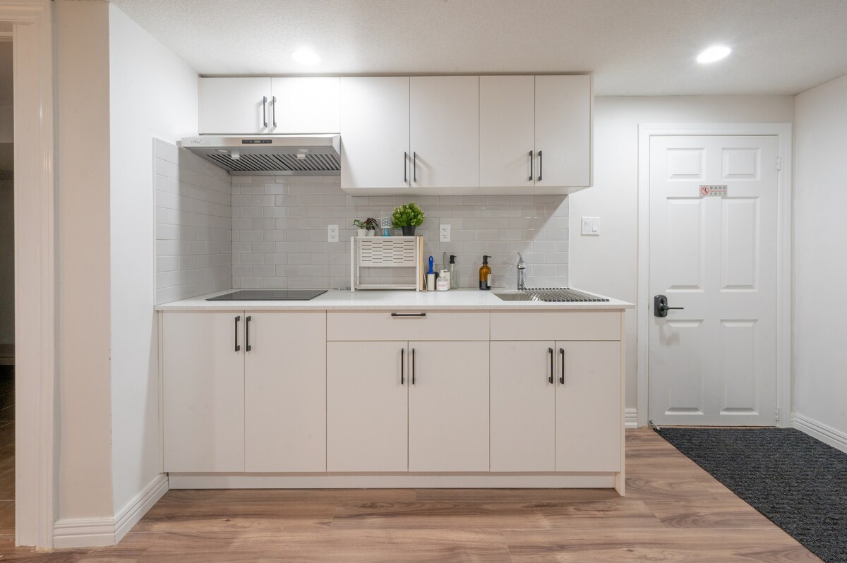 A modern kitchen is presented with sleek white cabinetry and a minimalist design. A stainless steel sink and stovetop are visible, accompanied by various kitchen supplies on the countertop. Soft lighting enhances the functional layout, while a door leads to an entryway.