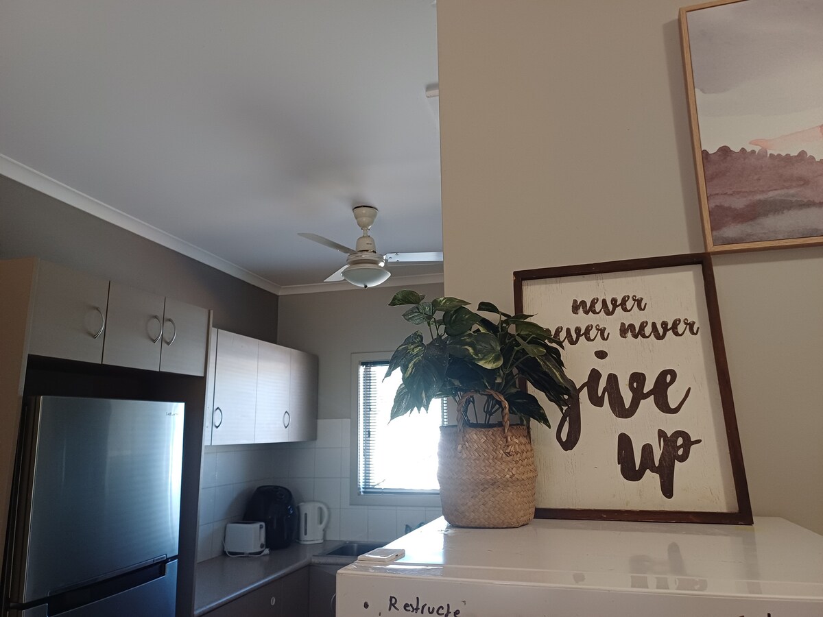 A kitchen area displays modern cabinetry and appliances, including a large refrigerator. A potted plant sits atop the refrigerator beside a framed motivational quote. Natural light enters through a window, enhancing the functional and welcoming environment.