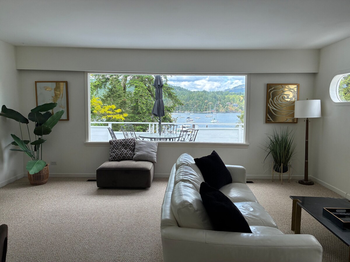 A spacious living area is highlighted by a large window offering views of the serene waterfront. A neutral-colored sectional couch and a decorative armchair complement the modern decor. A small dining table with chairs is positioned on the balcony, framed by greenery.