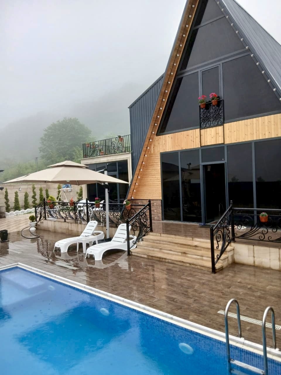 An A-frame villa with a wooden exterior is featured, surrounded by a misty atmosphere. A swimming pool sits in front, with white lounge chairs positioned nearby. Large glass doors provide views of the interior, while planters with flowers adorn the upper balcony.