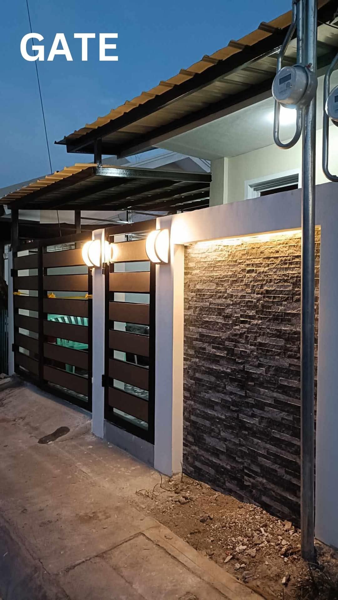 A modern entrance gate features horizontal wooden slats and a stone accent wall, illuminated by two round wall lights. The combination of materials creates a welcoming entryway. The surrounding area is paved, providing a clean approach to the property.