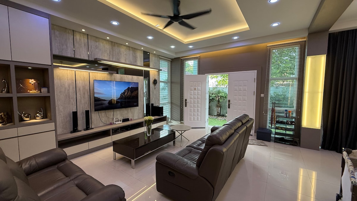 The spacious living area features a large flat-screen TV mounted on a textured wall. Two leather sofas are arranged around a sleek coffee table, while natural light enters through large windows. The adjoining glass doors lead to an outdoor space, enhancing openness.