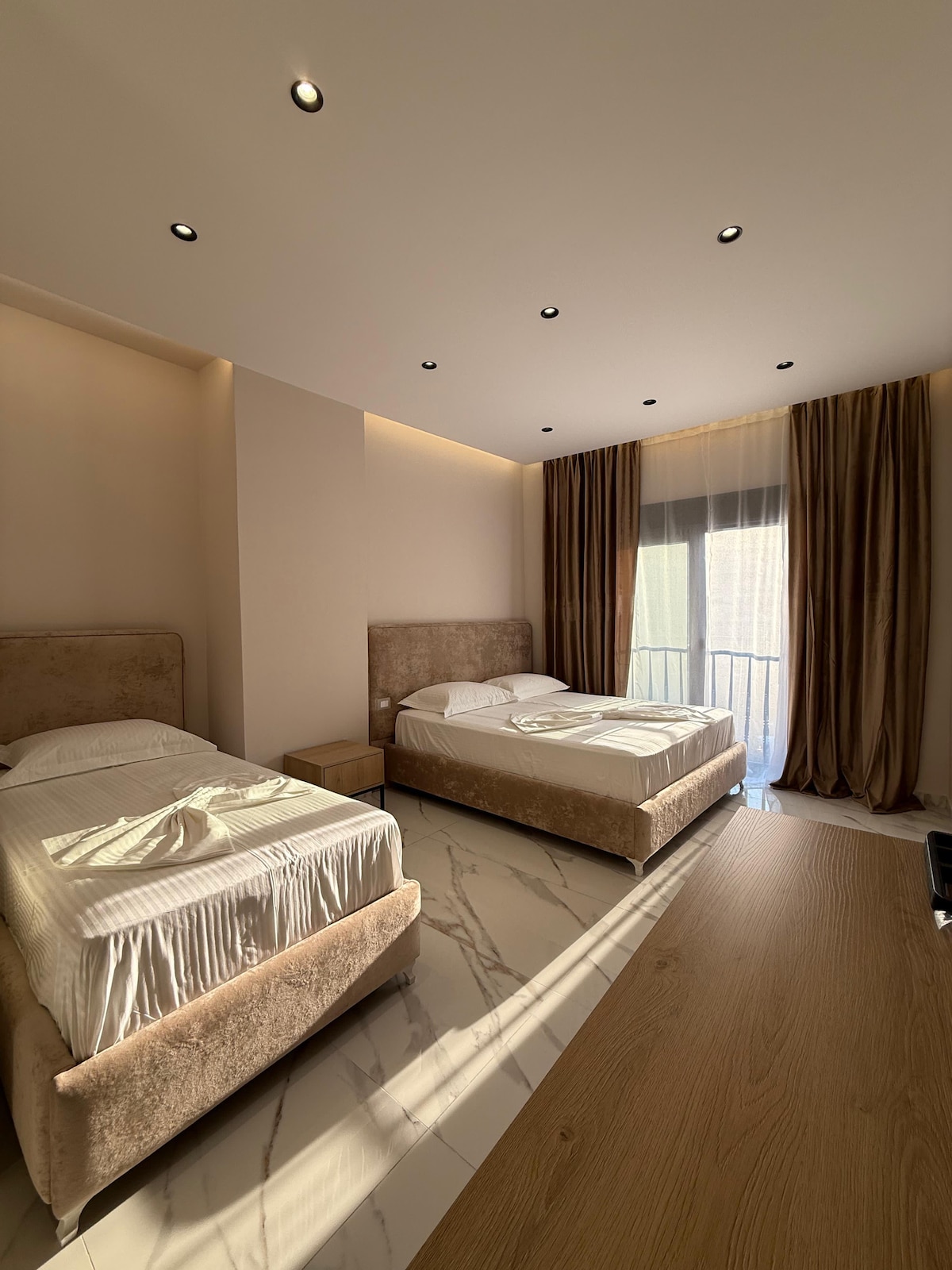 The room features two spacious beds, dressed in neutral linens. Soft, natural light filters through the window, creating gentle shadows on the floor. A wooden dresser complements the light-colored decor, while floor-to-ceiling curtains frame the window, offering privacy and elegance.
