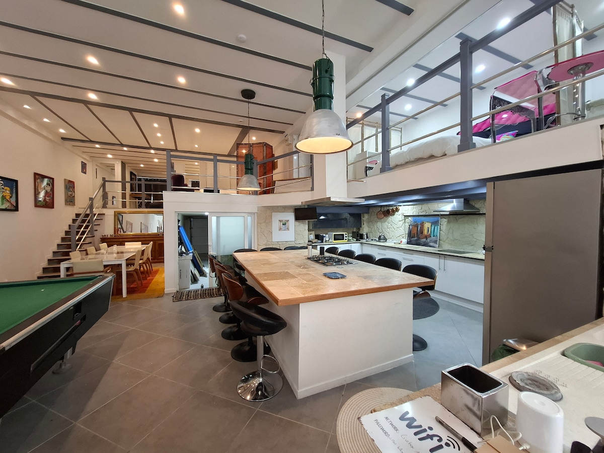 A spacious loft interior is depicted, featuring an expansive kitchen island surrounded by modern bar stools. A pool table is visible in the foreground, while the upper level with multiple sleeping areas is accessible via a staircase, showcasing a vibrant artful atmosphere.