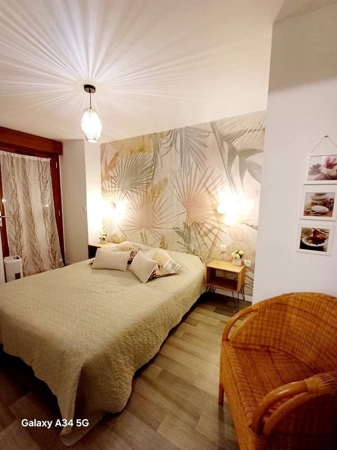 A cozy bedroom features a spacious bed dressed in neutral tones, flanked by warm wall lamps. A decorative wall with a botanical design adds a touch of nature. A rattan chair sits beside a small bedside table, enhancing the inviting atmosphere of the space.