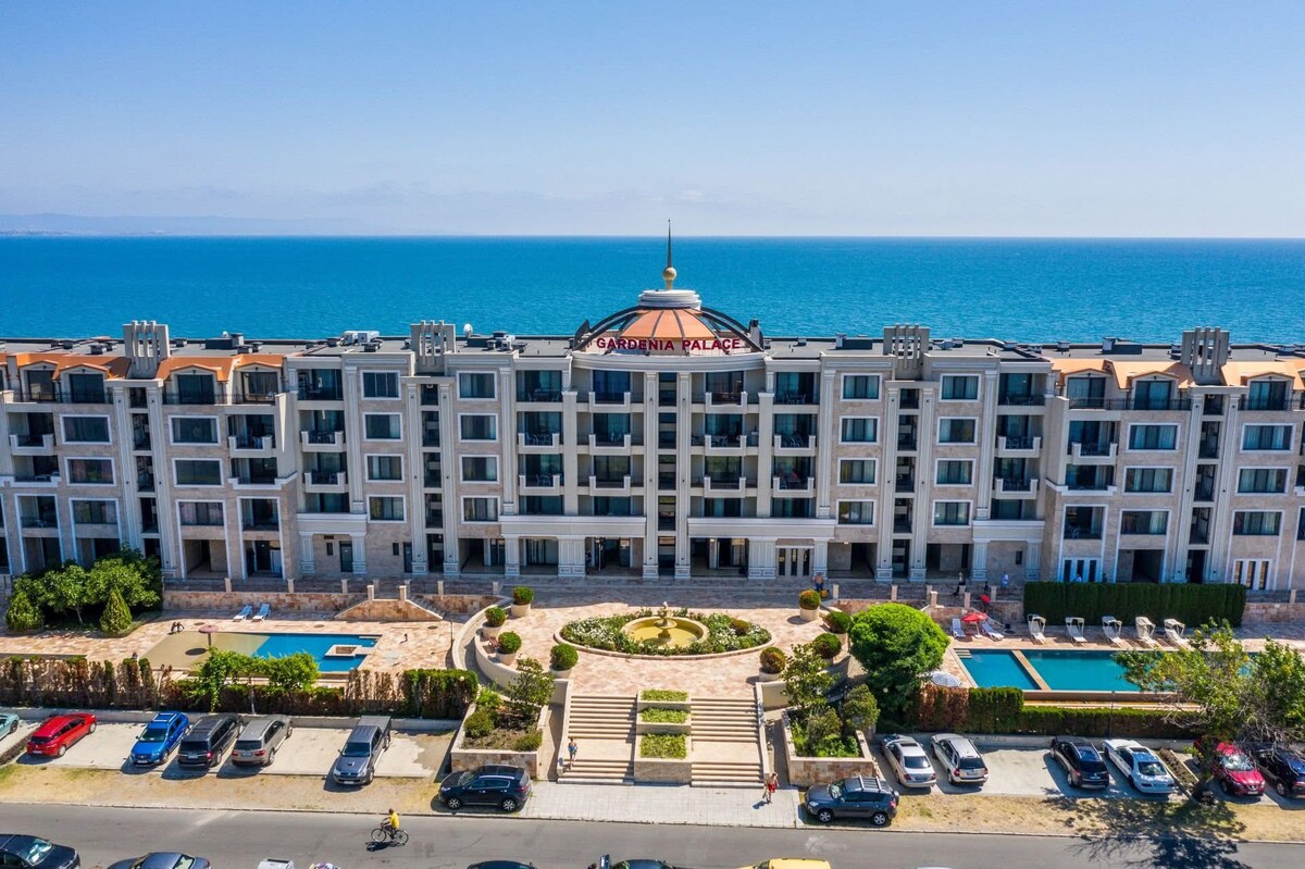 The beachfront apartment complex features a modern architectural style, with a signature dome at the center. The building is framed by vibrant landscaping and two outdoor pools. The expansive ocean is visible in the background, complementing the resort-like atmosphere of the property.