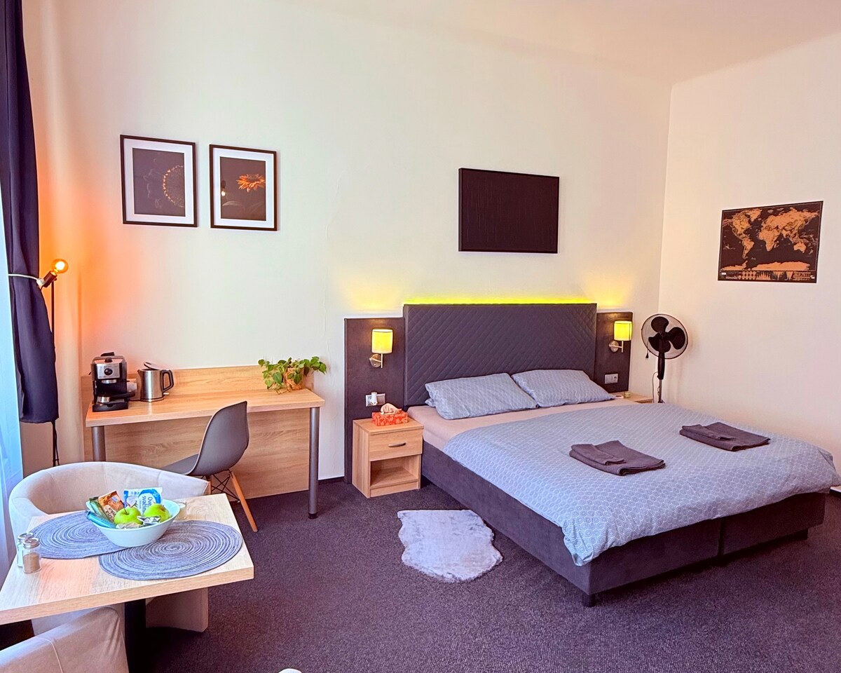 A cozy studio space features a spacious bed with neutral linens, flanked by two bedside lamps. A small table with a kettle and coffee supplies is positioned next to a dining area. Wall art in the form of framed photographs and a world map contribute to the inviting decor.