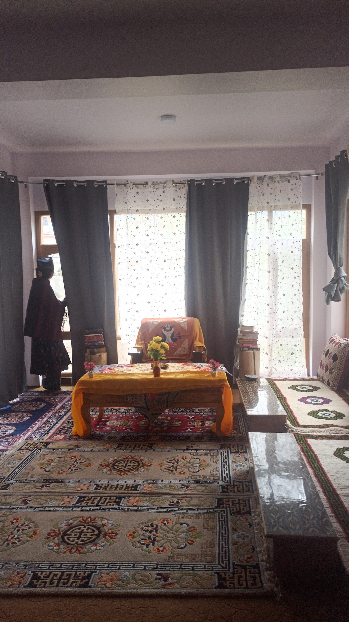 A cozy living area is featured, with large windows covered by sheer and dark curtains. A colorful patterned carpet covers the floor. A low table sits at the center, surrounded by comfortable seating and decorative items, creating an inviting space for relaxation.