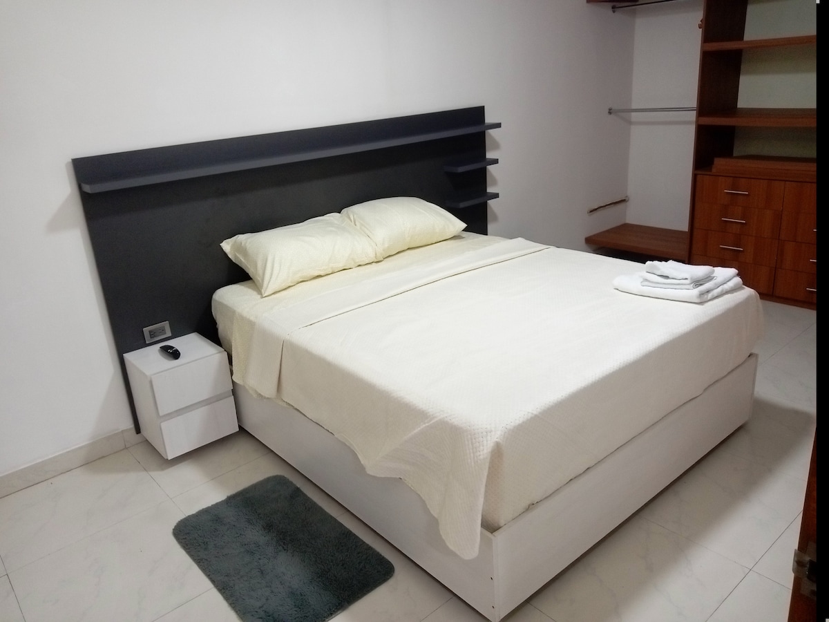 A spacious bedroom features a large bed with light-colored bedding. A nightstand is positioned beside the bed, equipped with a lamp. Shelves are mounted on the wall above the bed, and the room has a modern closet space along with a wooden dresser visible in the background.
