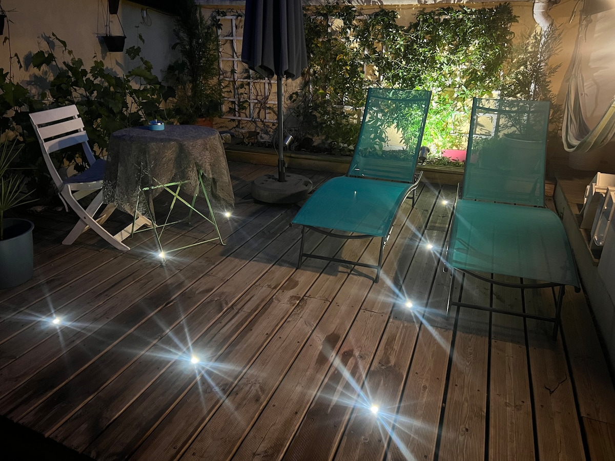 An outdoor terrace features two lounge chairs in soft blue, complemented by a round table set with a decorative cloth. The wooden decking has embedded lights, creating a gentle illumination, while greenery provides a natural backdrop for relaxation.