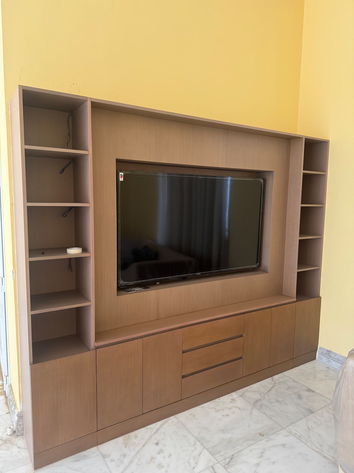 A modern media unit is displayed against a yellow wall, featuring a large flat-screen television at its center. Shelves are positioned on either side, providing space for decorative items. The unit is complemented by a lower cabinet with multiple drawers for storage.