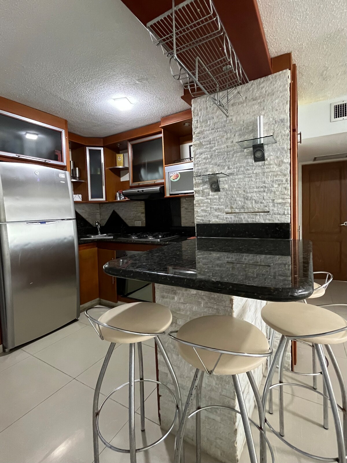 The kitchen features sleek cabinetry and a dark countertop, complemented by a light-colored stone wall. Three modern bar stools are positioned around a small breakfast bar, while stainless steel appliances, including a refrigerator, are visible in the background.