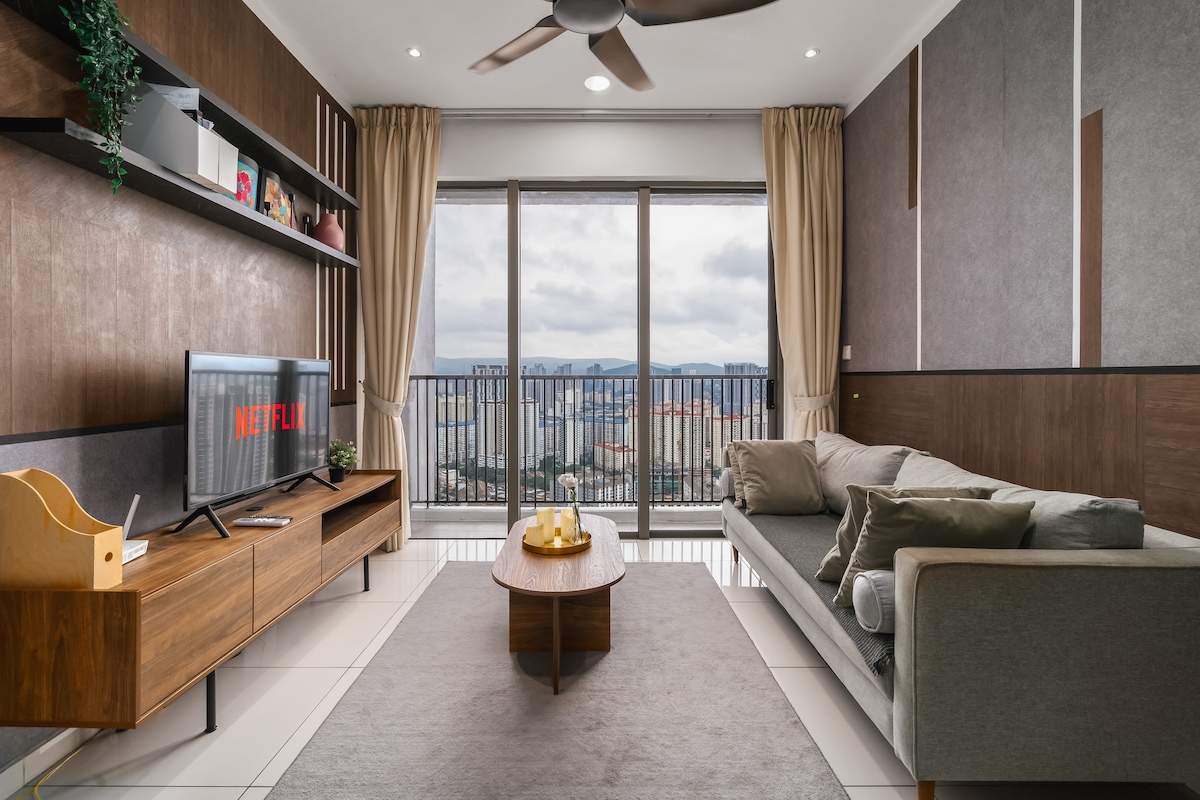 The living area features a comfortable sofa arranged around a low coffee table. A wall-mounted TV with a streaming device is visible, along with large windows offering expansive views of the city skyline. Light-colored curtains and a ceiling fan provide a bright, airy ambiance.