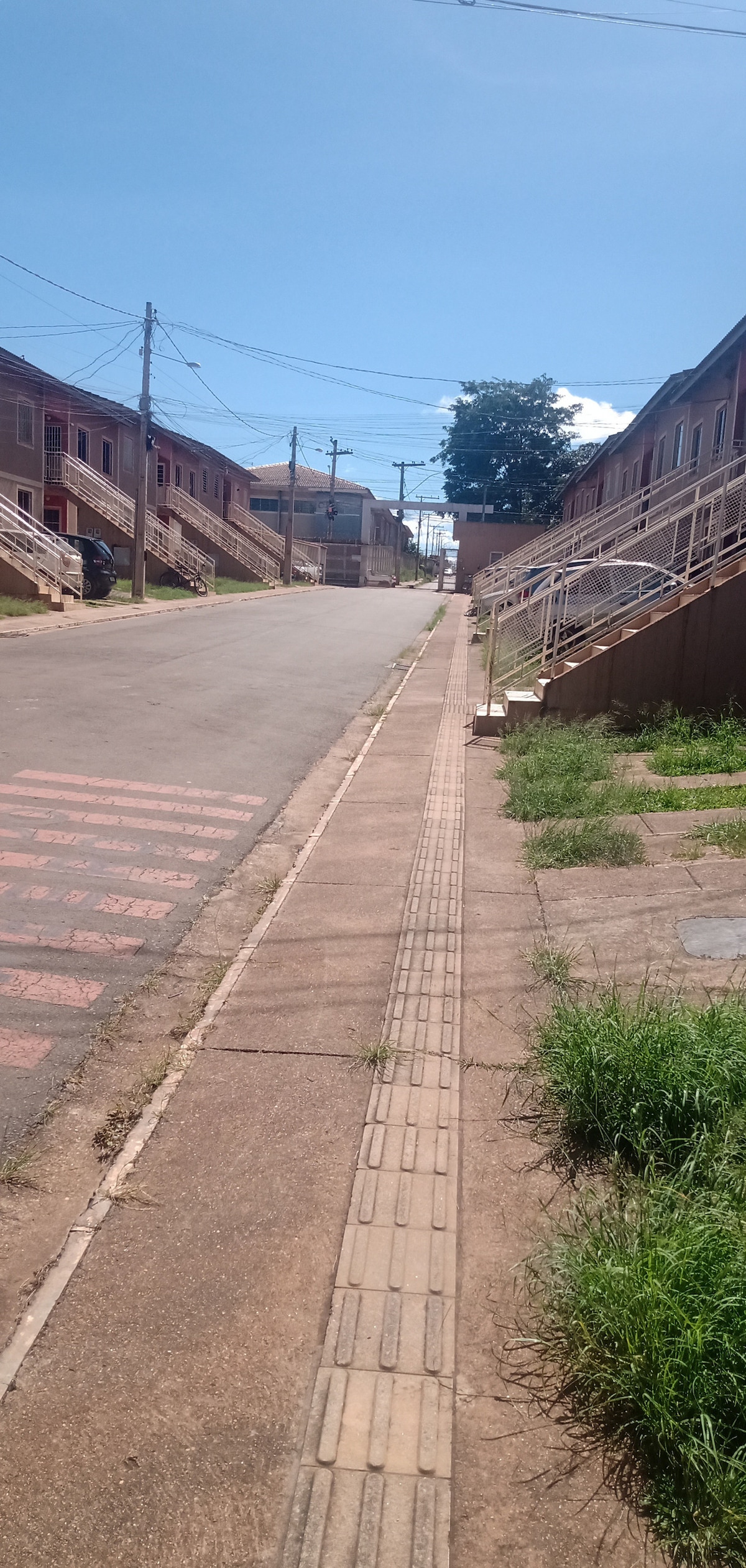 A wide, sunlit street is depicted, lined with modest buildings on either side. The gently sloping sidewalk features tactile paving for guidance. Grass grows alongside the curb, while utility poles and power lines are visible in the clear blue sky.