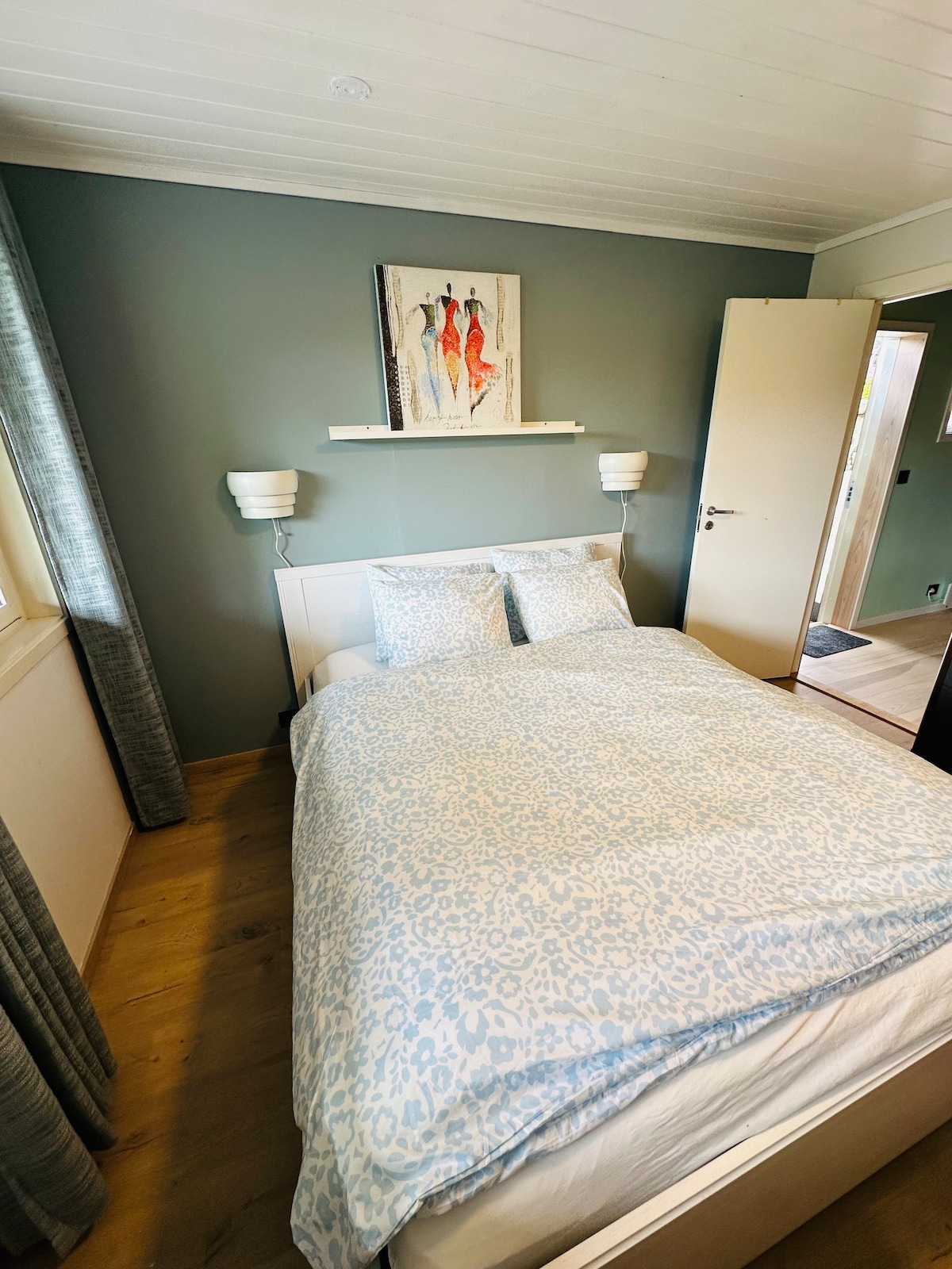 A neatly arranged sleeping area is presented, featuring a spacious bed adorned with a light blue patterned duvet. Soft lighting is provided by wall-mounted lamps on either side. A decorative artwork hangs above the bed, and a partially open door reveals a glimpse of the hallway.