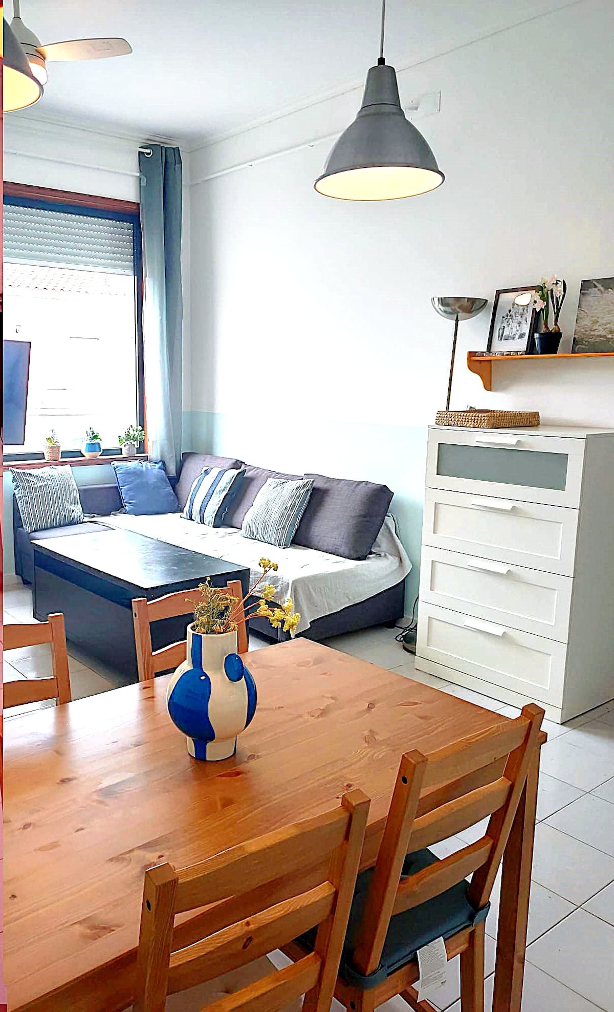 A cozy living area is depicted, featuring a comfortable sofa bed with neutral-colored cushions. A dining table for six is positioned nearby, complemented by wooden chairs. Natural light enters through a large window, brightening the space and highlighting decorative items on shelves.