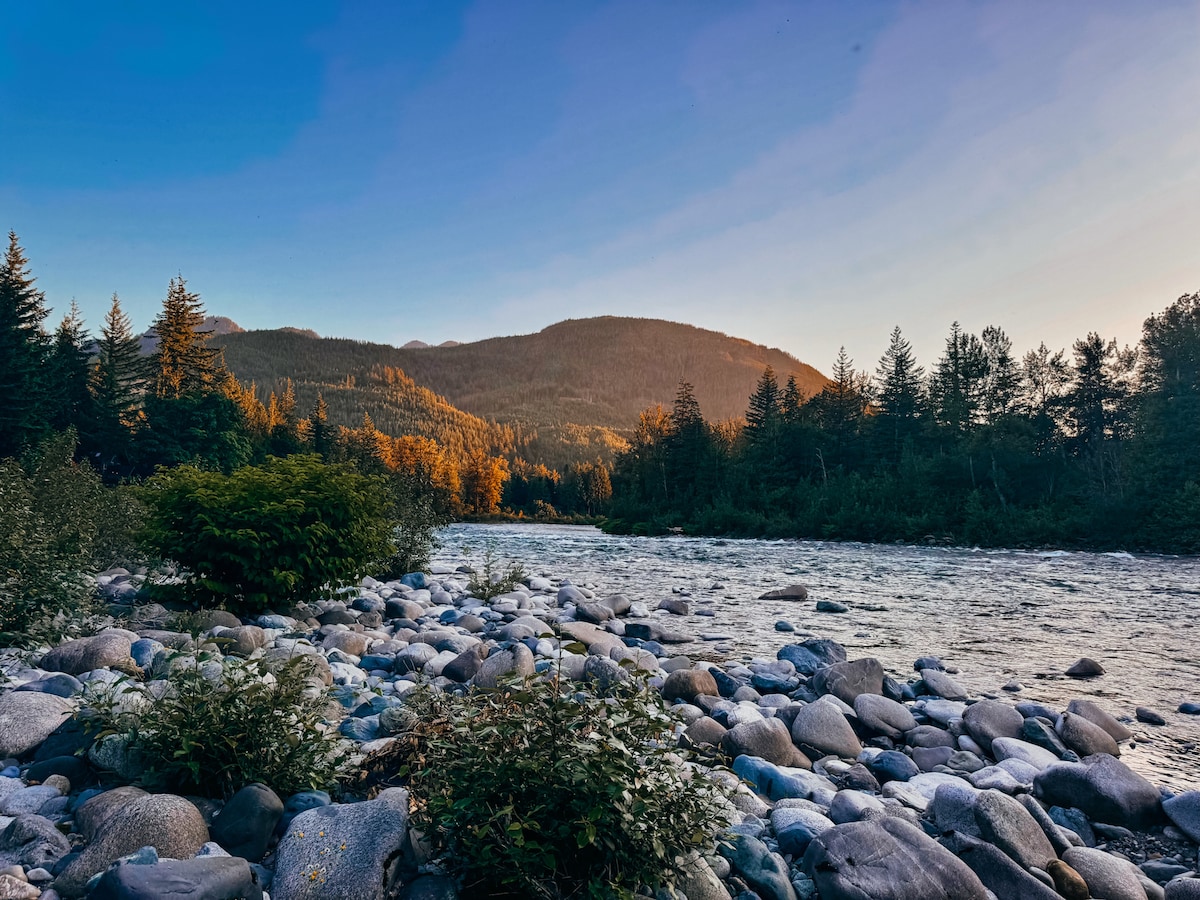 A rocky riverbank is framed by a gently flowing Skykomish River and lush greenery. Golden light illuminates the trees on the opposing hillside, while the distant mountains rise against a clear blue sky, creating a serene natural landscape.