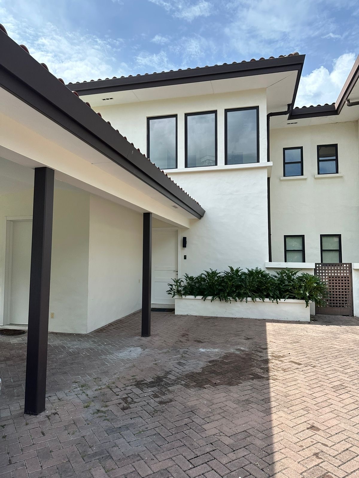 The exterior of a modern building features clean lines and a combination of white stucco walls and dark accents. A covered entrance is visible, framed by greenery, while patterned brick pavers lead towards the structure, reflecting a welcoming environment.