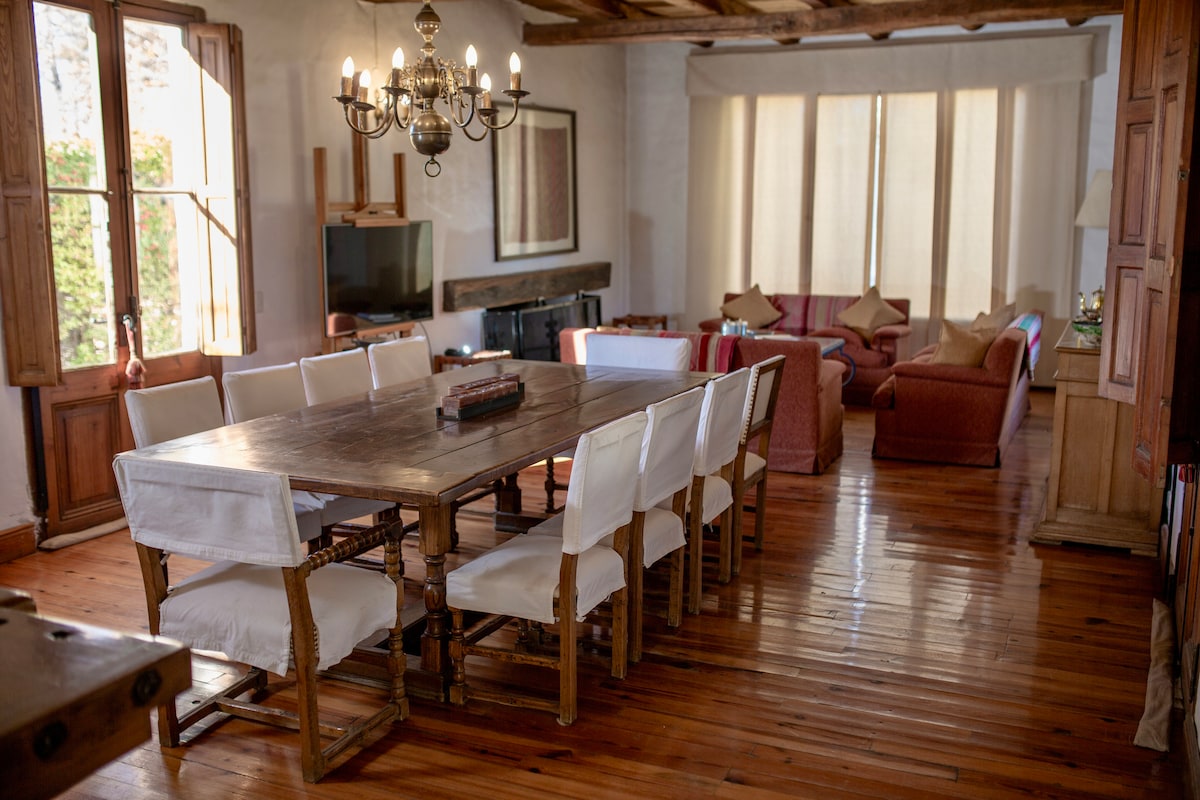 A large wooden dining table is surrounded by ten white chairs, positioned in a spacious dining area. Natural light filters through large windows, illuminating the warm wooden floor and inviting decor. A cozy seating arrangement with red sofas is visible in the background.